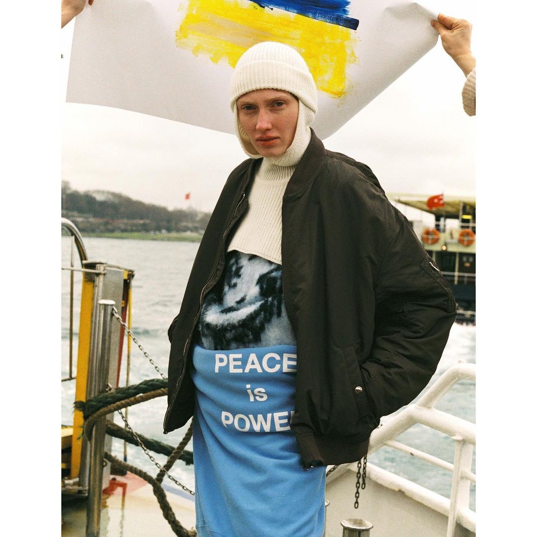 Person holding a Ukrainian flag with painted colors, wearing a white hat and dark coat, stands on a boat with water visible behind.