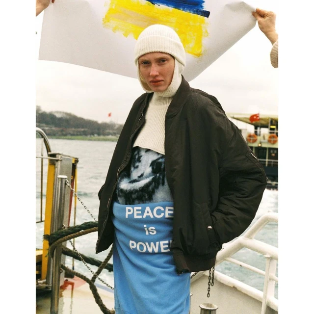 Person holding a Ukrainian flag with painted colors, wearing a white hat and dark coat, stands on a boat with water visible behind.