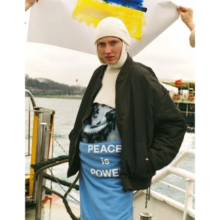 Person holding a Ukrainian flag with painted colors, wearing a white hat and dark coat, stands on a boat with water visible behind.