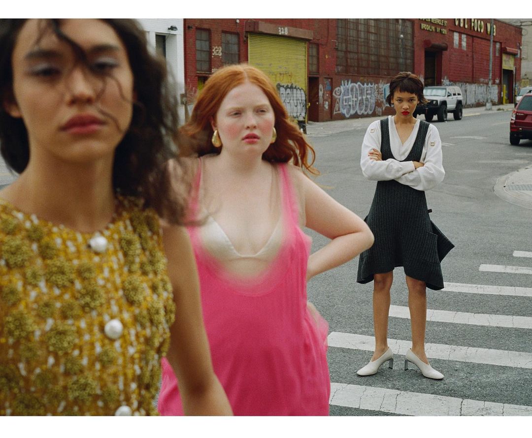 Three women stand on a crosswalk in a city street. One wears a yellow top, one a pink dress, and one a gray dress with a white shirt.