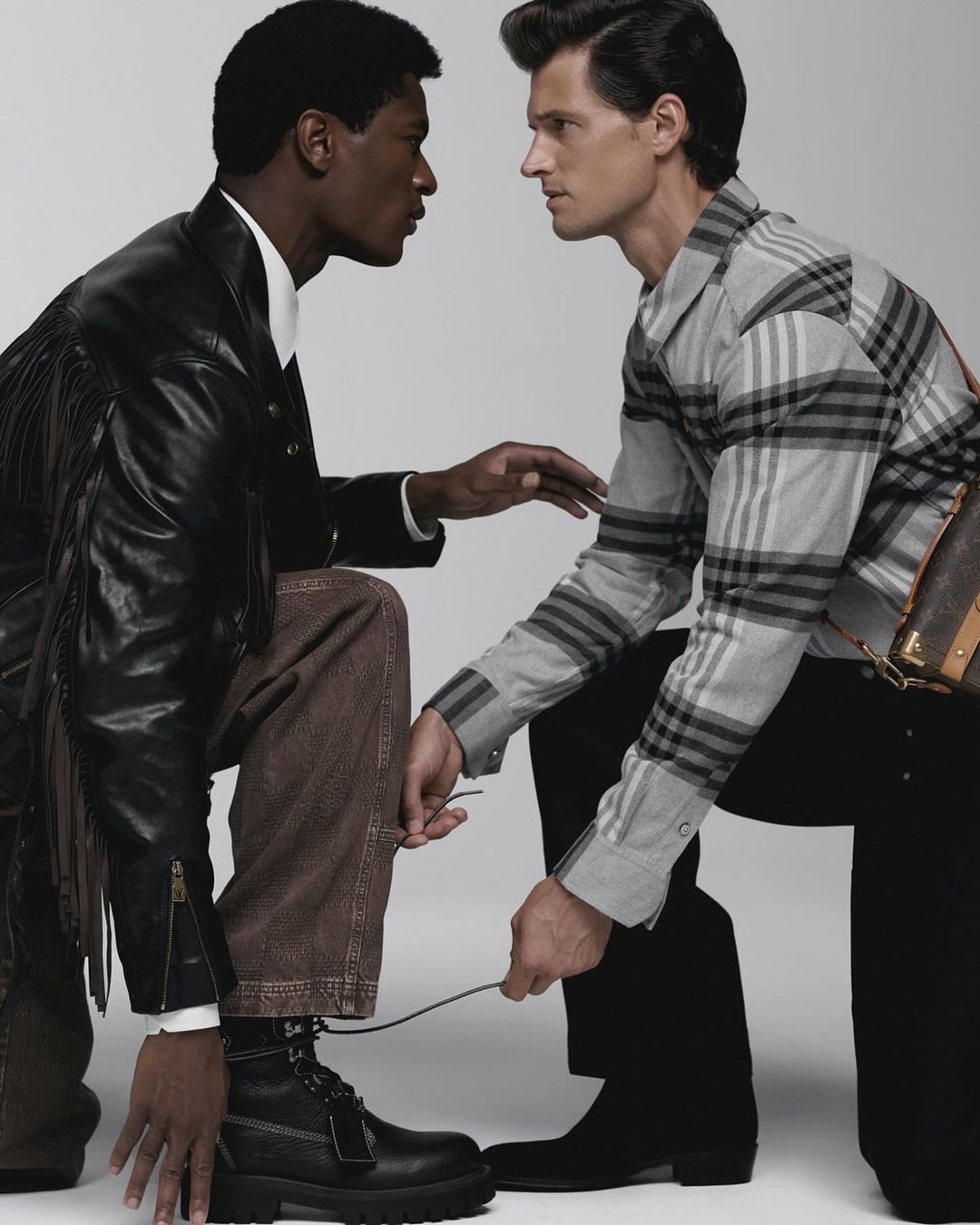 Two men in a fashion shot sitting together, one tying the other's boot. Dark leather jacket, jeans, plaid shirt, boots, and a neutral background.