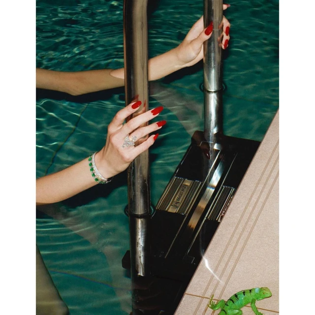 Woman's hands with red nails and jewelry grip a pool ladder in clear water, a toy lizard sits on the pool deck nearby.