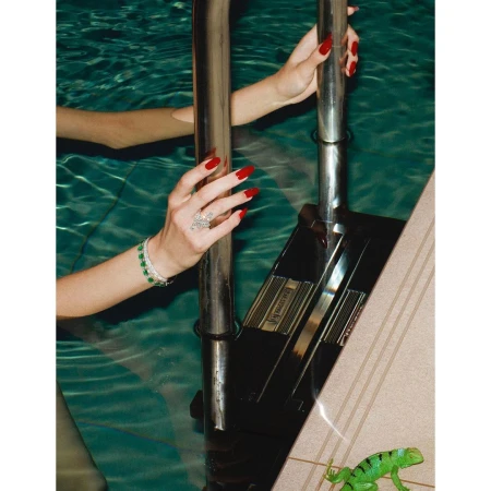 Woman's hands with red nails and jewelry grip a pool ladder in clear water, a toy lizard sits on the pool deck nearby.