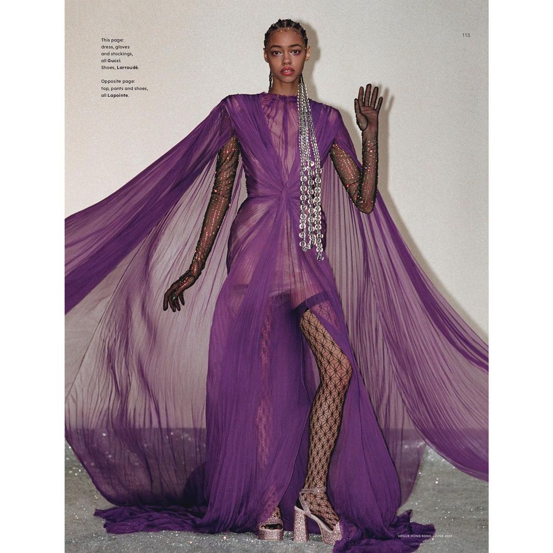 Fashion editorial shot of a woman in a purple gown with a flowing cape, black gloves, patterned stockings, and embellished platform heels.
