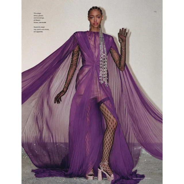Fashion editorial shot of a woman in a purple gown with a flowing cape, black gloves, patterned stockings, and embellished platform heels.