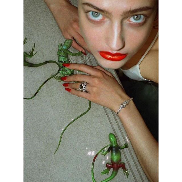 Adult female with striking blue eyes and red lips poses with toy green lizards on a light tile surface, wearing jewelry.