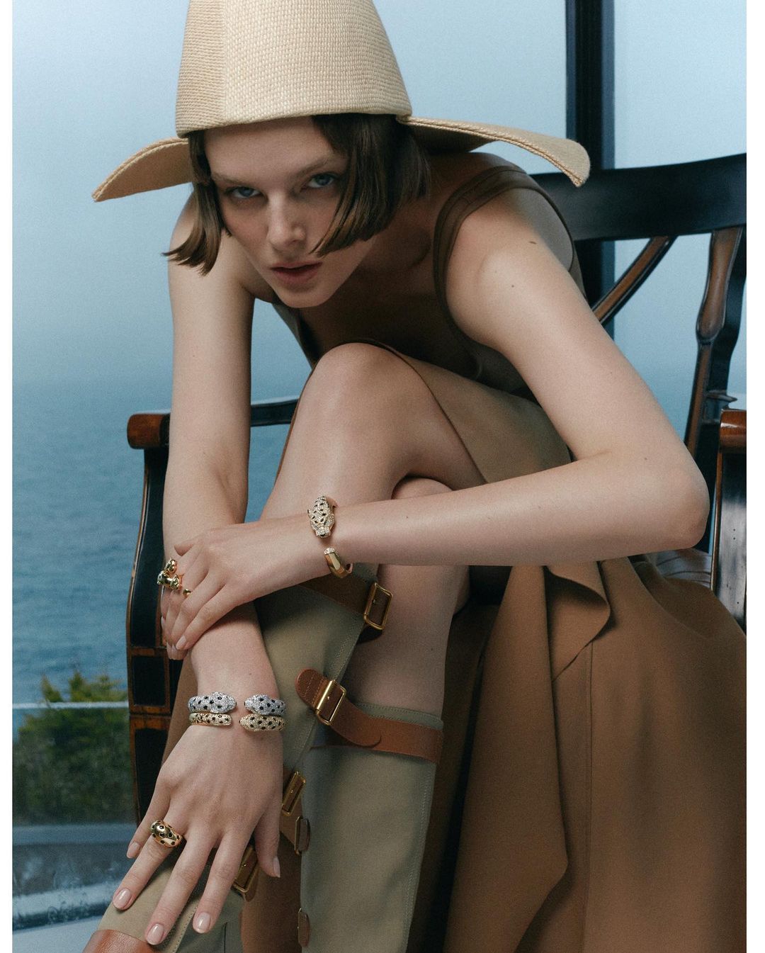 Brunette woman in a hat, khaki dress, and boots, sitting in front of a window overlooking the ocean while wearing rings and bracelets.