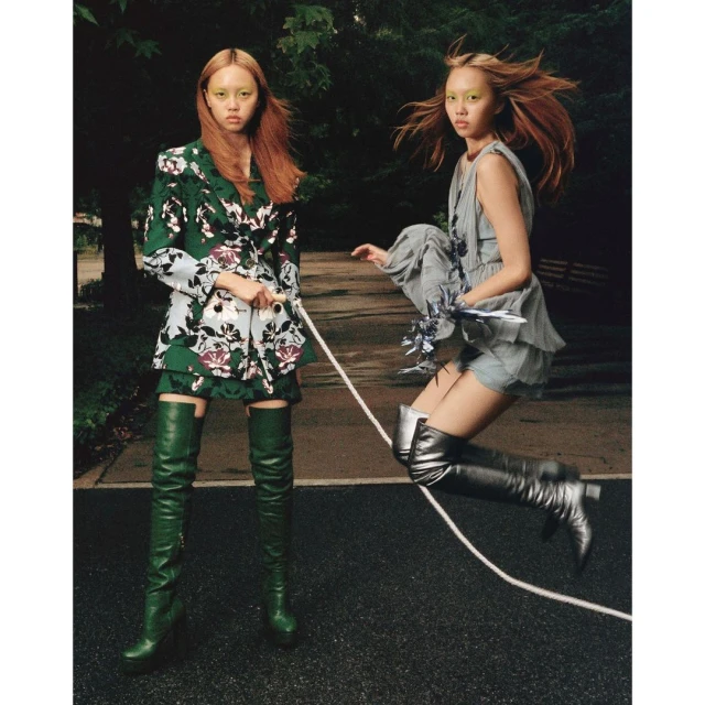 Two fashion models pose outdoors in colorful outfits with floral designs, one jumping in the air while holding a white rope, wearing silver thigh-high boots.