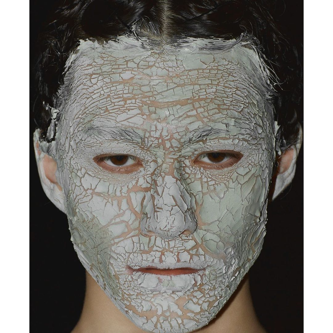 Close-up of a person's face covered in a cracked, pale-green clay mask against a dark background, showcasing beauty or skincare treatment.