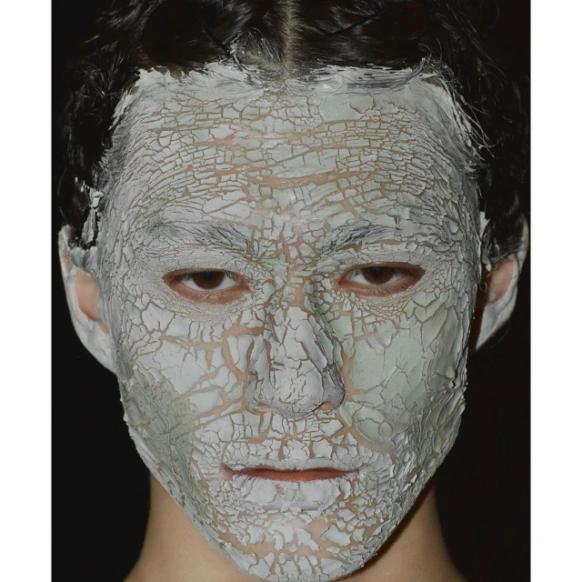 Close-up of a person's face covered in a cracked, pale-green clay mask against a dark background, showcasing beauty or skincare treatment.