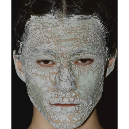 Close-up of a person's face covered in a cracked, pale-green clay mask against a dark background, showcasing beauty or skincare treatment.