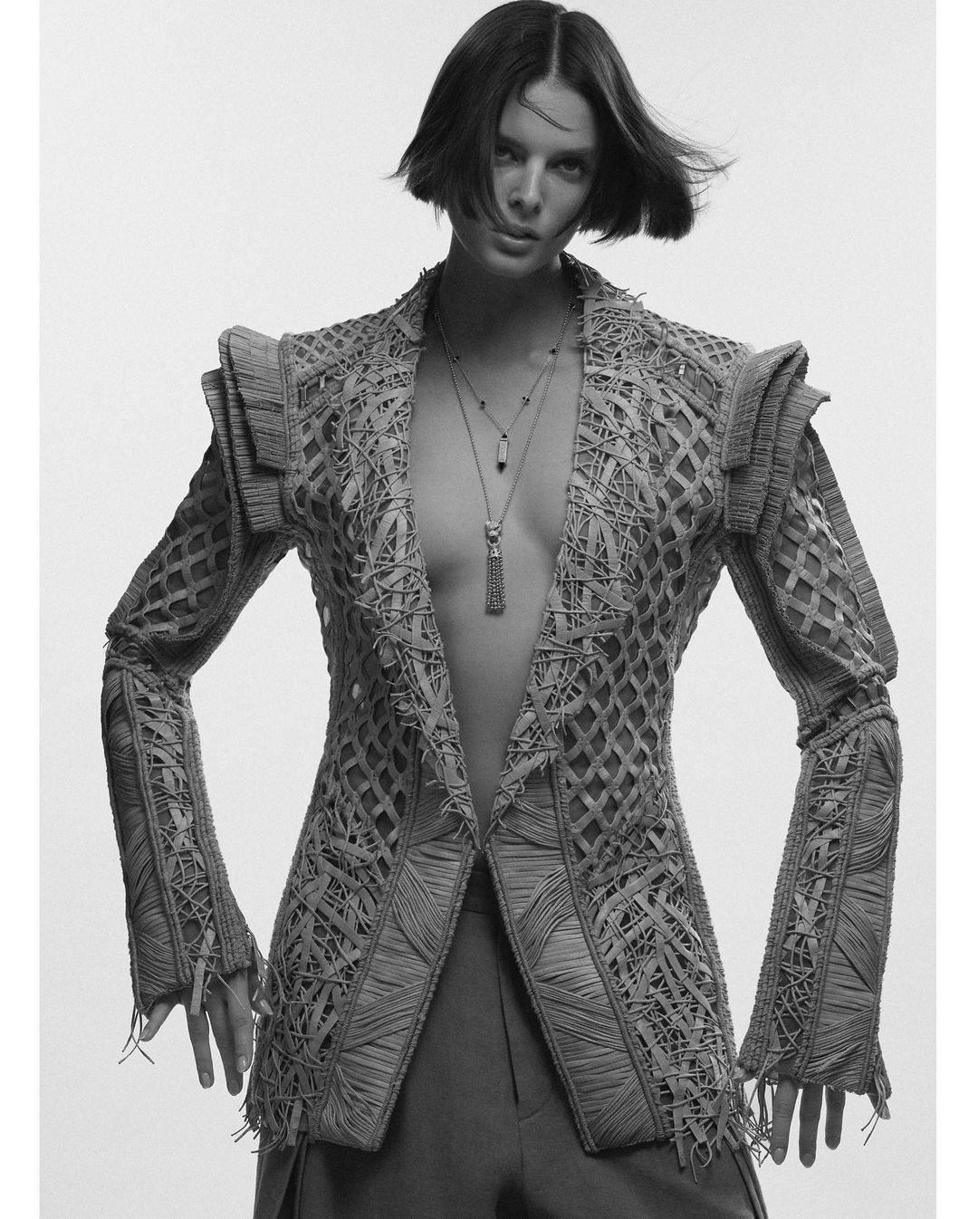 Monochrome portrait of a woman wearing a tailored jacket with detailed weaving and multiple pendant necklaces against a white backdrop.
