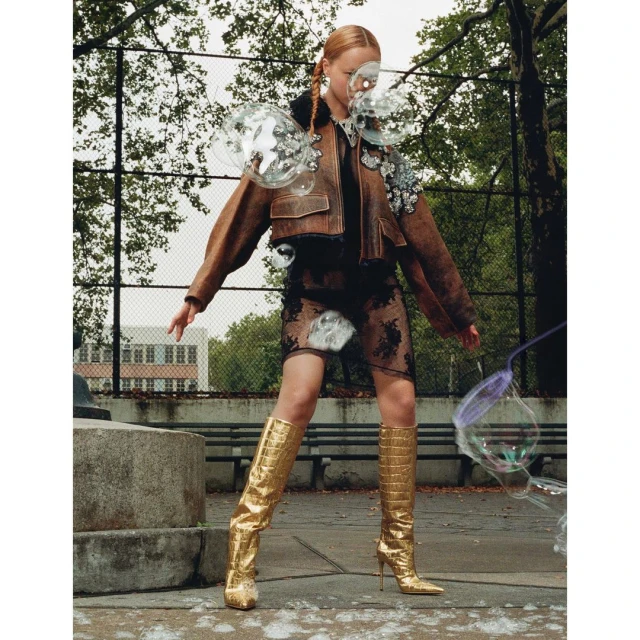 Fashion shot of a woman in a brown jacket and gold boots standing in an urban park with bubbles, building and trees in the background.