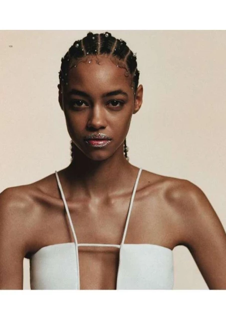 Close-up portrait of a young African-American woman with decorative braids wearing a white top, set against a neutral-toned background.
