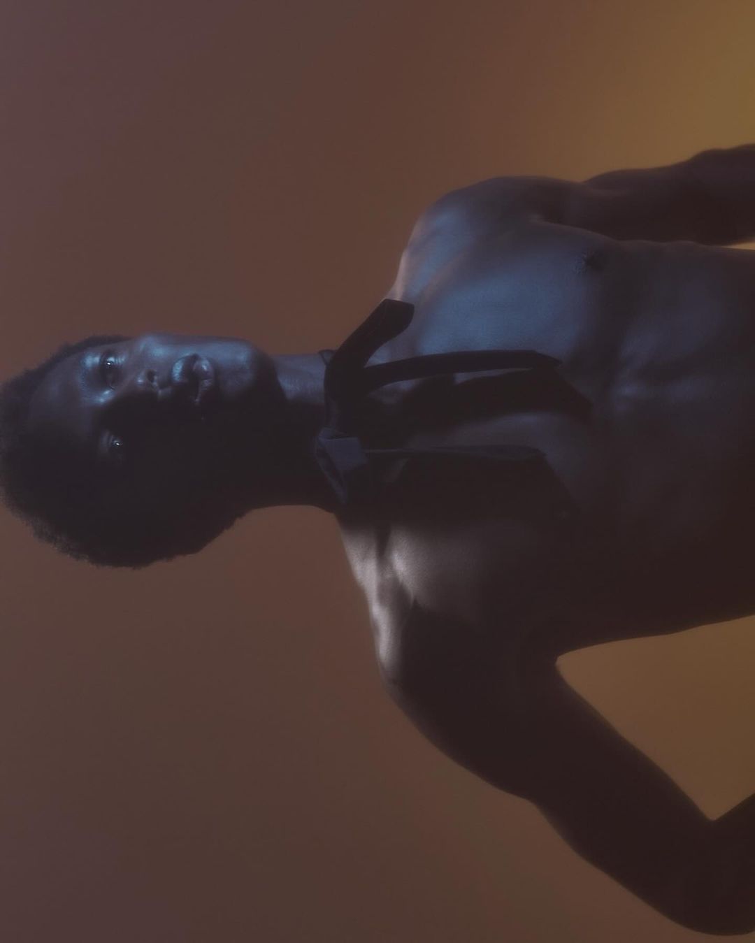 Portrait of a Black man with a muscular build, captured in a dark and moody atmosphere with warm yellow and orange lighting.