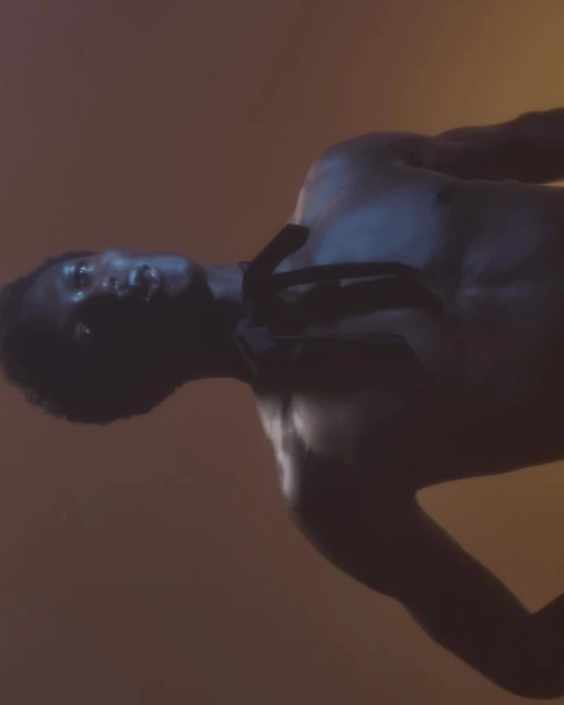 Portrait of a Black man with a muscular build, captured in a dark and moody atmosphere with warm yellow and orange lighting.