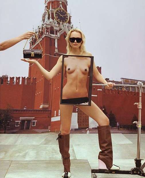 Fashion shot of a woman in boots holding a clear frame and handbag with Red Square in the background, posing on a rooftop stage.
