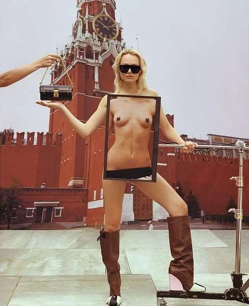 Fashion shot of a woman in boots holding a clear frame and handbag with Red Square in the background, posing on a rooftop stage.