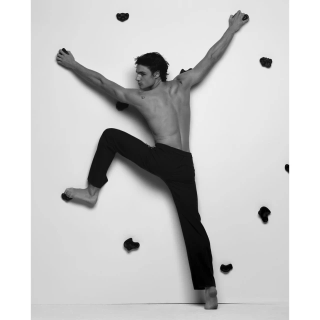A man in black pants climbing a white wall with hand and foot holds, arms outstretched, captured in a black and white studio shot.
