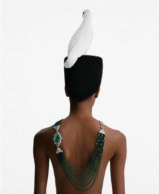 Back view of androgynous model with emerald necklace and white bird on head against a white background in a studio lighting setup.