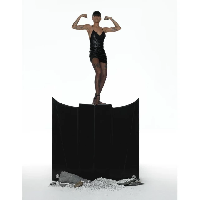 An adult female poses on a black abstract structure, flexing biceps against a bright white background in a studio photograph.