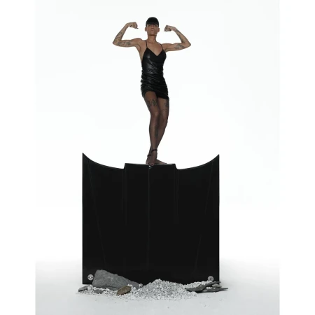 An adult female poses on a black abstract structure, flexing biceps against a bright white background in a studio photograph.