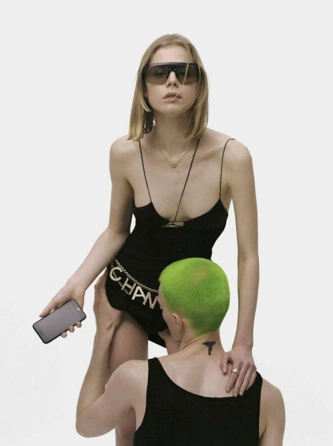 Two models in a photo studio. A female model in a black dress stands while a second model kneels with green hair. 