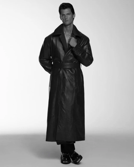 A man in a long leather coat strikes a pose in a monochromatic photo with a simple studio backdrop. He is wearing black shoes.