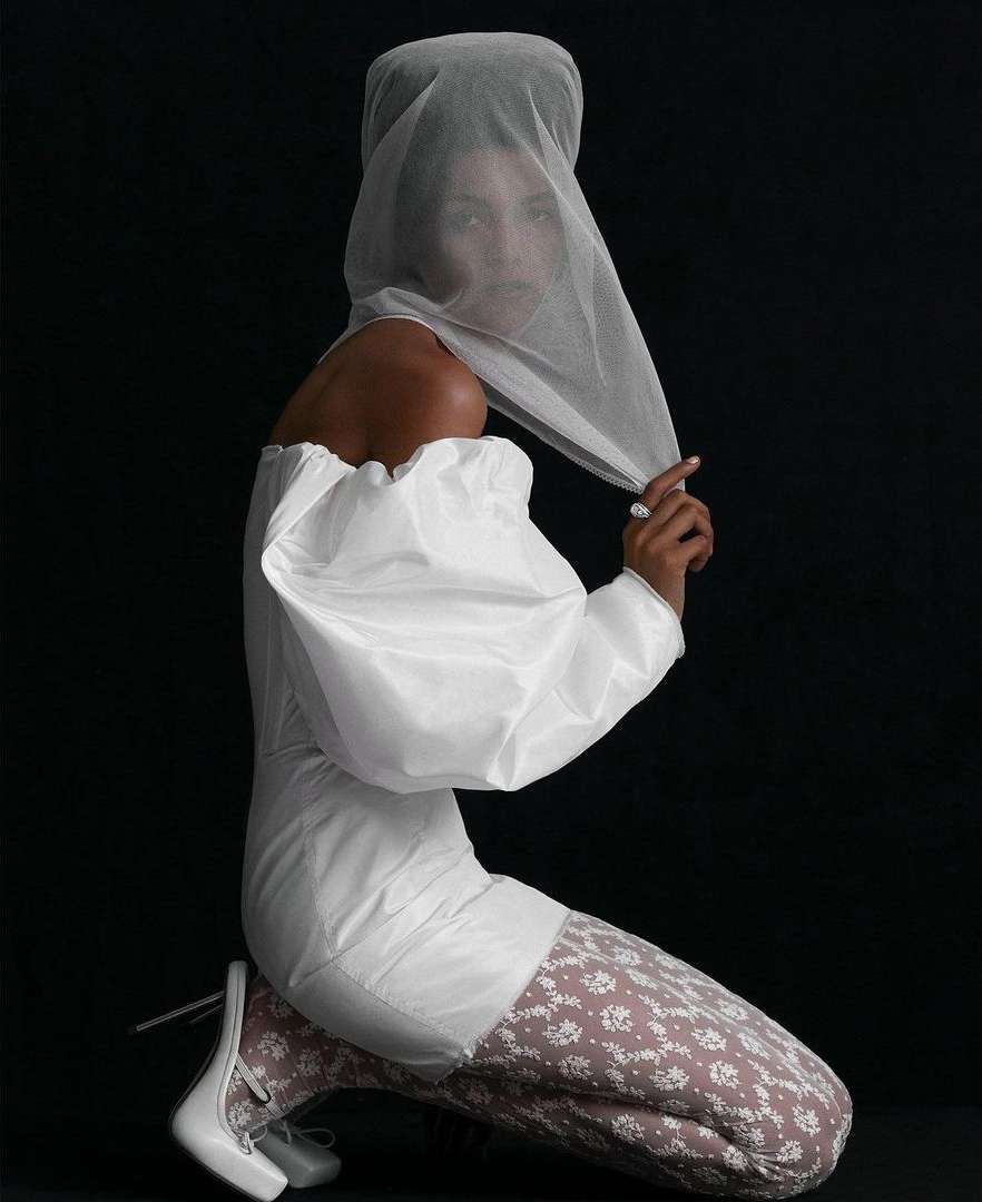 Woman poses kneeling in a studio wearing an off-shoulder white dress, patterned leggings, white shoes, and a sheer head covering against black.