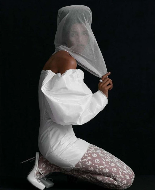 Woman poses kneeling in a studio wearing an off-shoulder white dress, patterned leggings, white shoes, and a sheer head covering against black.