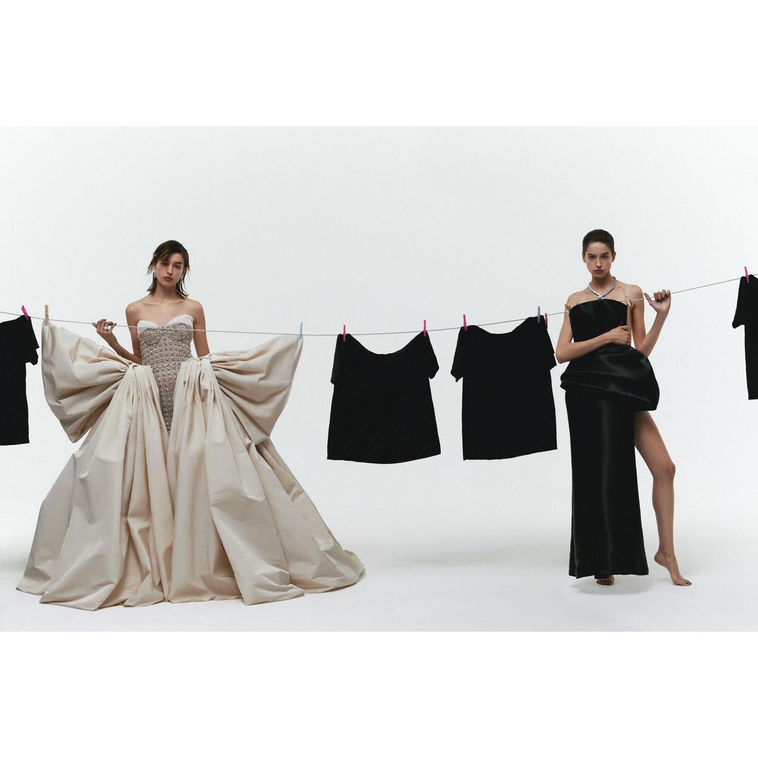 Two women pose in high fashion designs beside a clothesline with dark garments against a white backdrop in a studio setting.