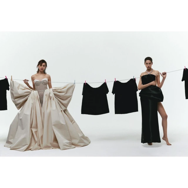Two women pose in high fashion designs beside a clothesline with dark garments against a white backdrop in a studio setting.