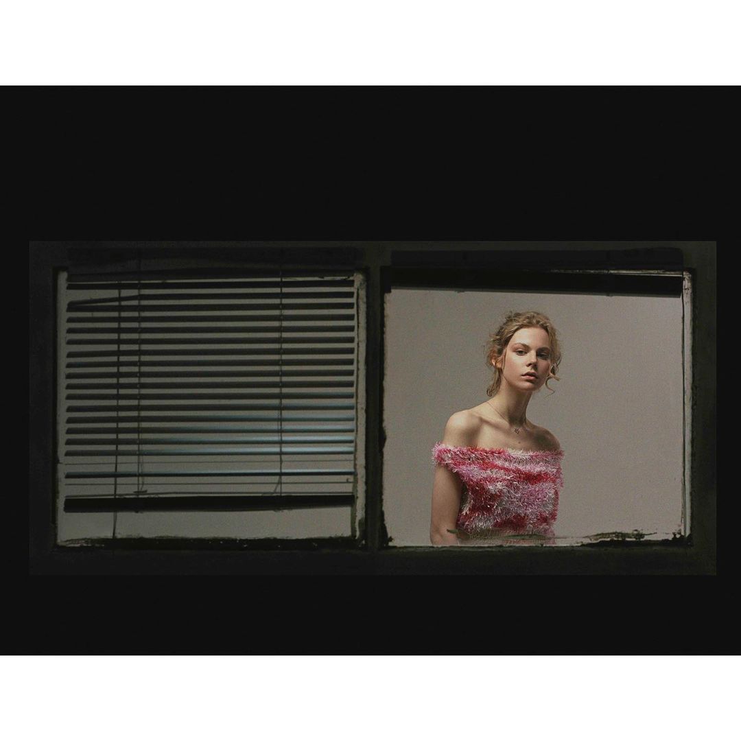 An eye-level portrait showcases a model framed by a window, featuring neutral walls, dark window frames, and light from window on the left.