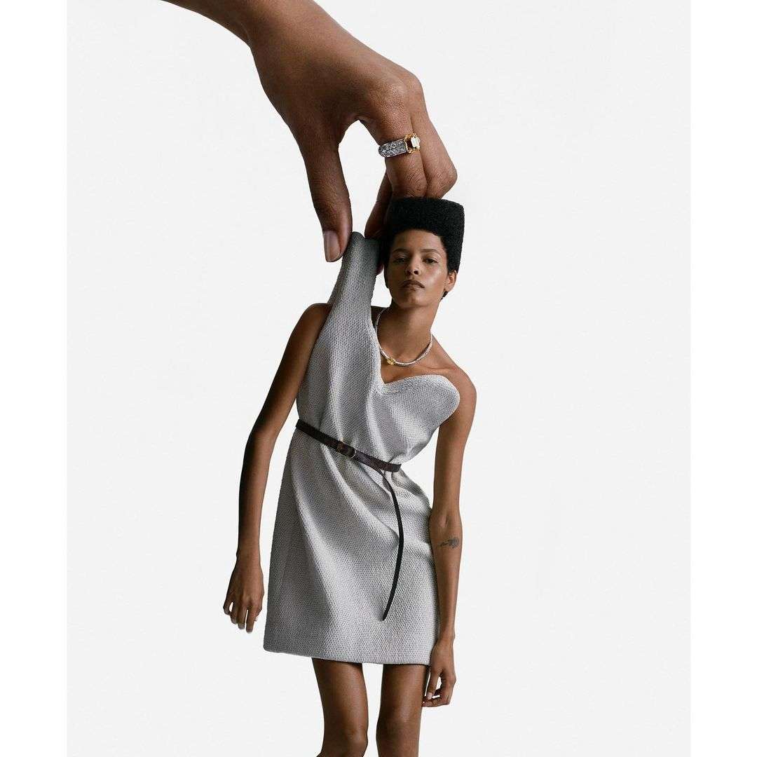 Fashion shot of a female model wearing a grey dress and belt, with an oversized hand holding her head in a white studio.