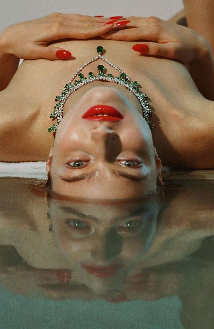 Adult female lying on her back with her hands above her chest wearing a green and white gemstone necklace and red lipstick and fingernails.