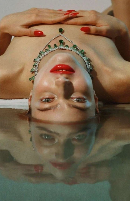Adult female lying on her back with her hands above her chest wearing a green and white gemstone necklace and red lipstick and fingernails.