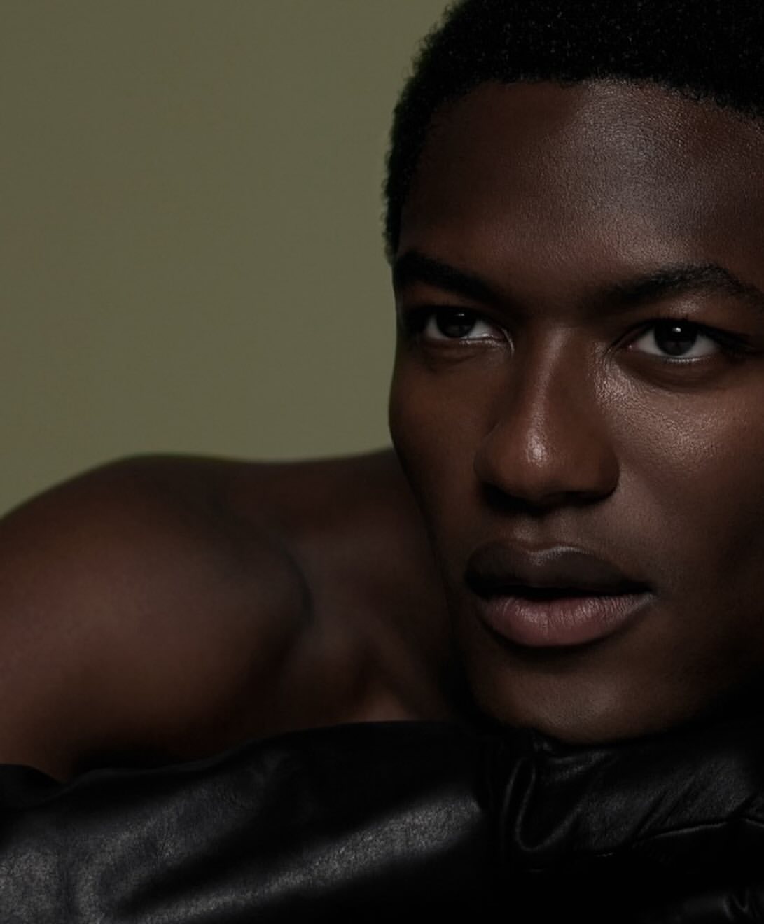 Close-up portrait of a young, dark-skinned, bare-chested man with short hair, looking off-camera and resting on black leather.