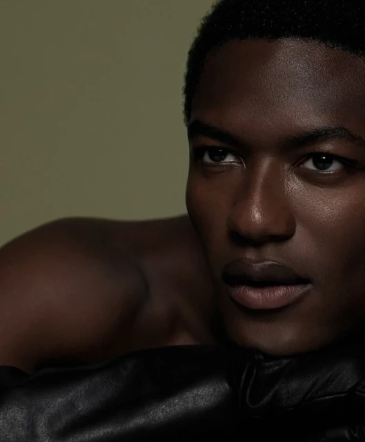 Close-up portrait of a young, dark-skinned, bare-chested man with short hair, looking off-camera and resting on black leather.