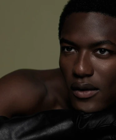 Close-up portrait of a young, dark-skinned, bare-chested man with short hair, looking off-camera and resting on black leather.