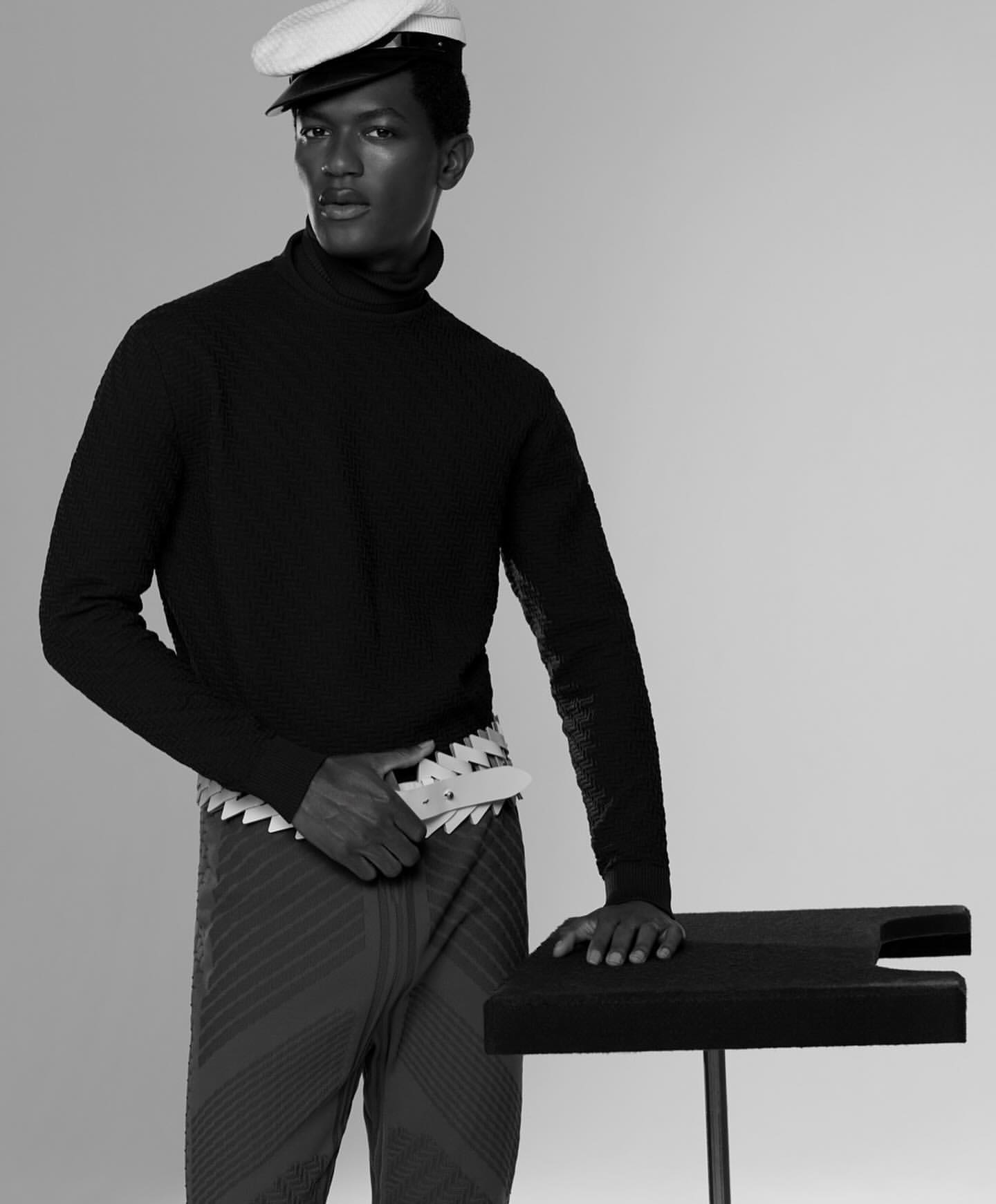 Monochrome studio shot of a Black male model in a turtleneck, patterned pants, and peaked cap, leaning on a black table. Fashion.