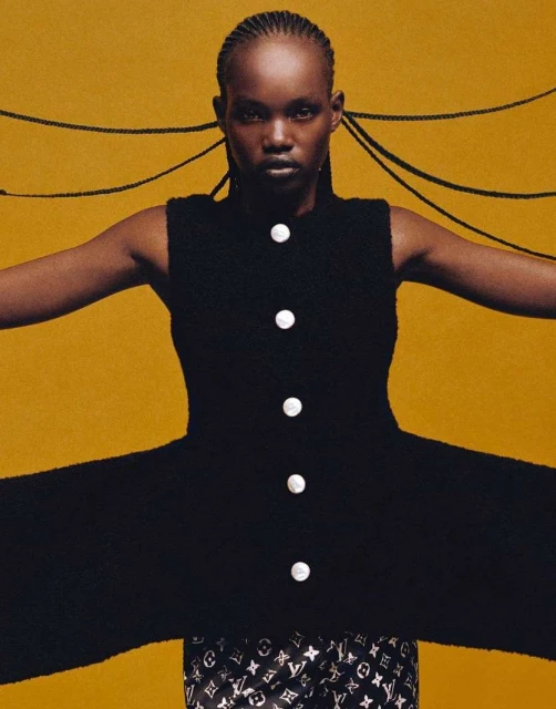 A woman with braids poses in a black dress with buttons against a yellow background, presenting a striking fashion and beauty portrait.