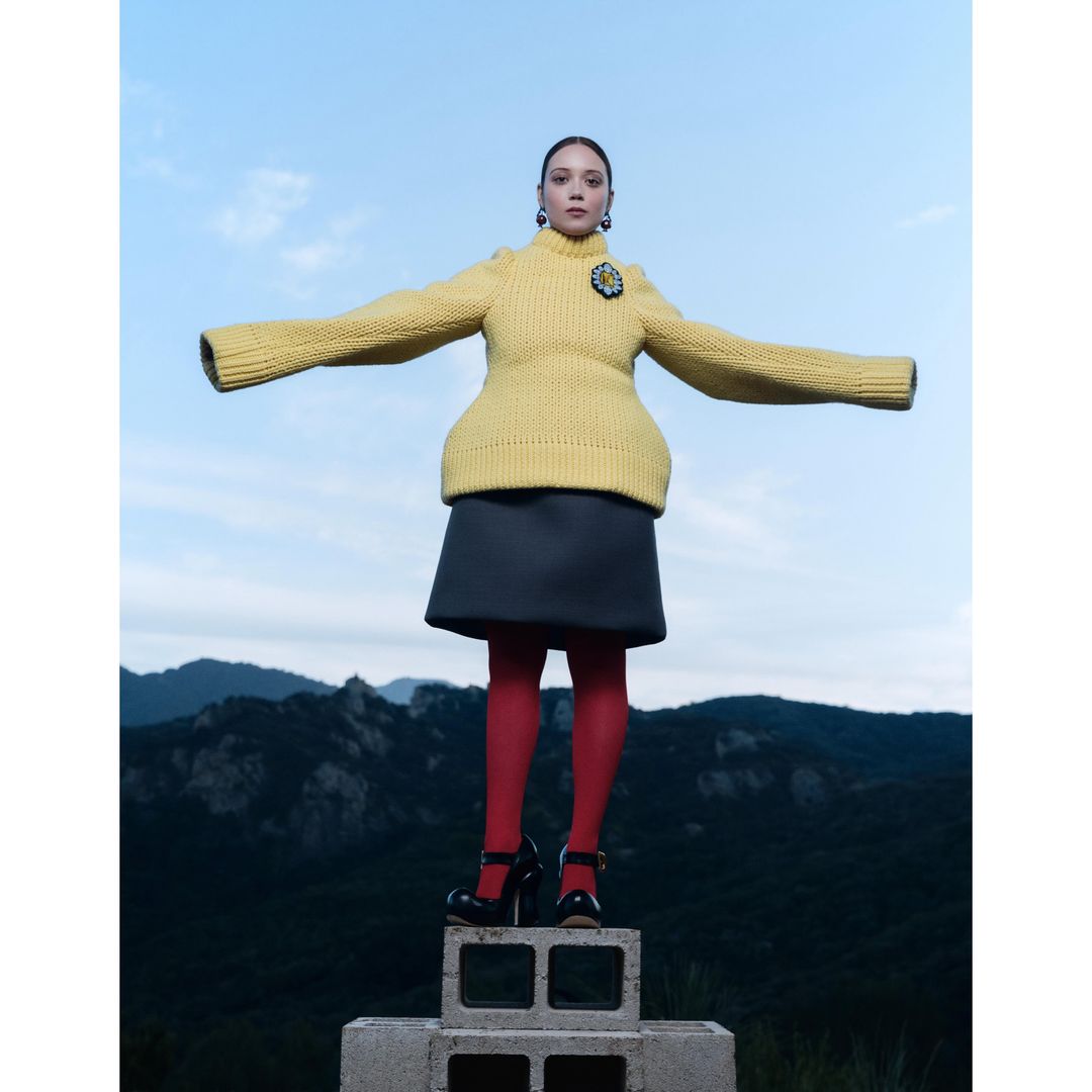 Woman in yellow sweater, grey skirt, red tights, and black shoes poses on concrete blocks against a mountain and sky background.