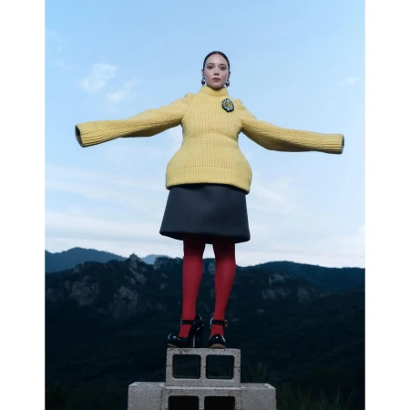 Woman in yellow sweater, grey skirt, red tights, and black shoes poses on concrete blocks against a mountain and sky background.