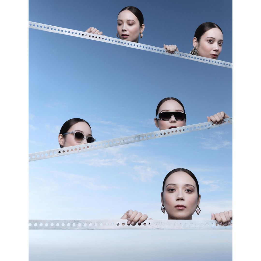 Fashion shot of a woman with multiple visible faces wearing sunglasses and jewelry, set against a blue sky, holding onto metal bars.
