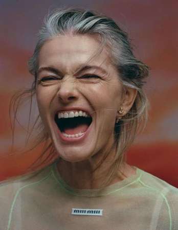 Close-up portrait of a woman with grey hair laughing hysterically in a sheer mint green top against an abstract gradient background.