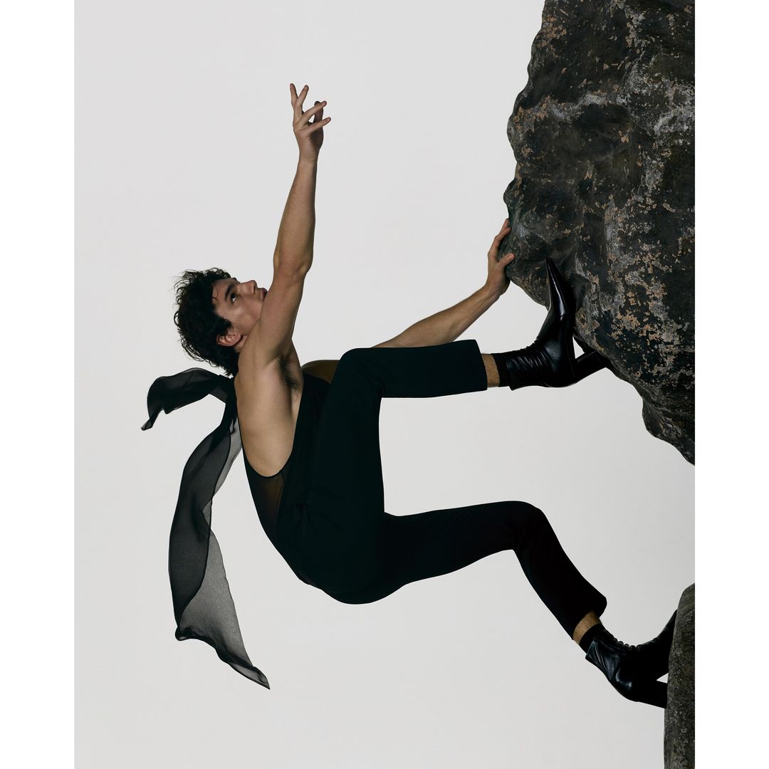 A man climbing a vertical rock face wearing a black sleeveless top, trousers, and boots with a flowing dark fabric.