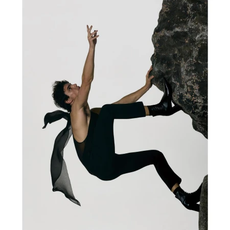 A man climbing a vertical rock face wearing a black sleeveless top, trousers, and boots with a flowing dark fabric.