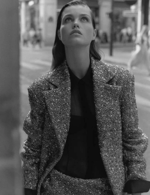Monochrome portrait of a woman wearing a tweed blazer and black top, looking up in an outdoor urban setting with soft light.