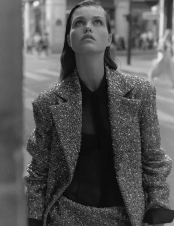 Monochrome portrait of a woman wearing a tweed blazer and black top, looking up in an outdoor urban setting with soft light.