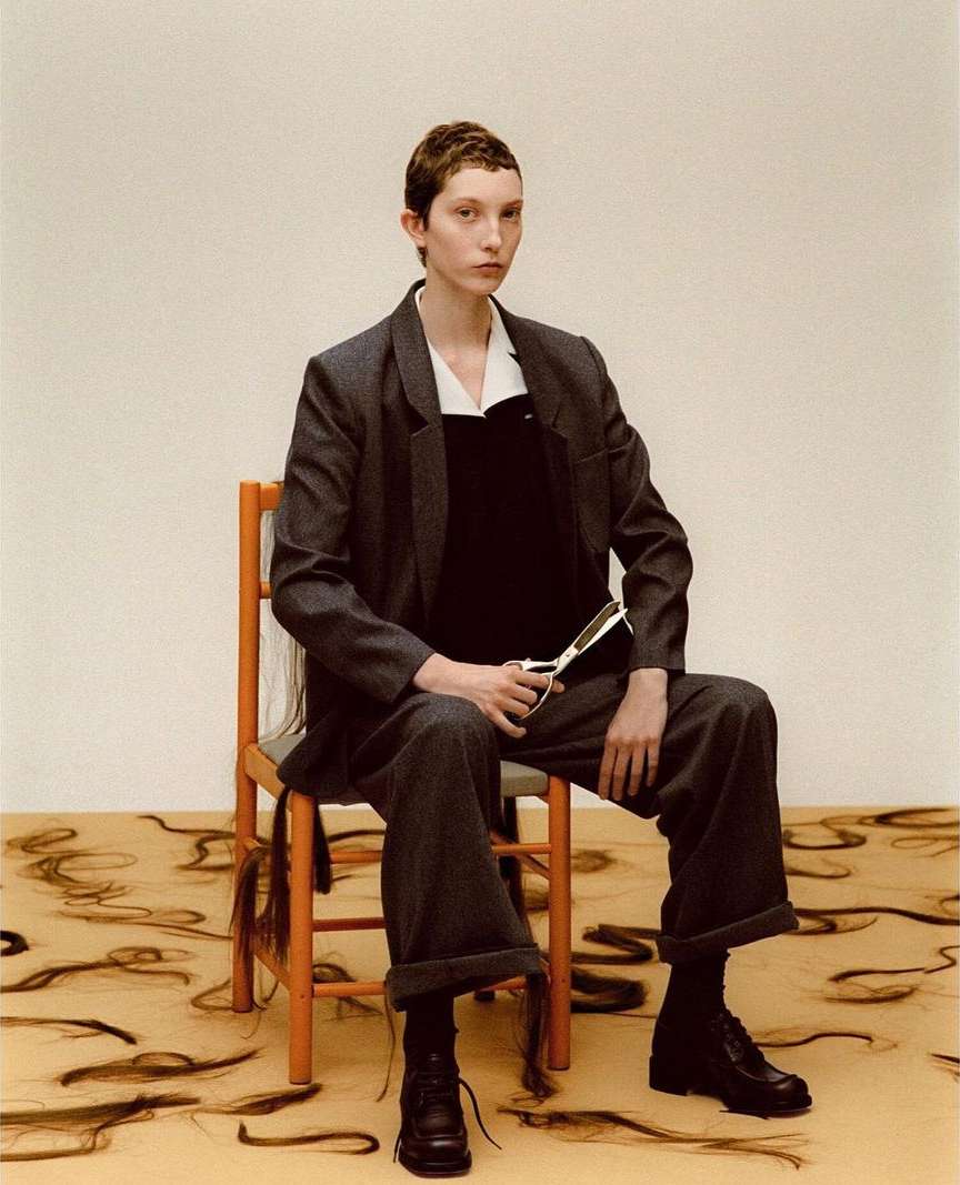 Model with short hair wearing a grey suit and holding scissors seated on an orange chair over scattered hair strands.
